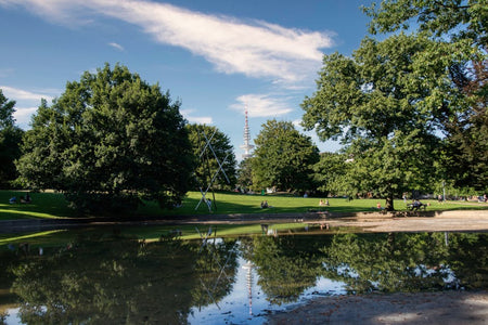 A tranquil park scene with lush trees, a reflective pond, and a distant tower under a sunny sky.