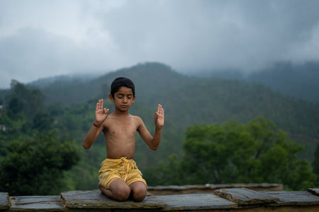 A young boy practices meditation outdoors with a peaceful mountain backdrop.