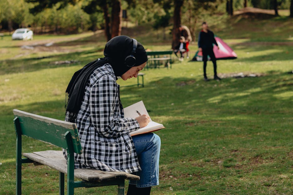 Woman in hijab writing in notebook on a bench in a sunny summer park.