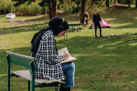 Woman in hijab writing in notebook on a bench in a sunny summer park.