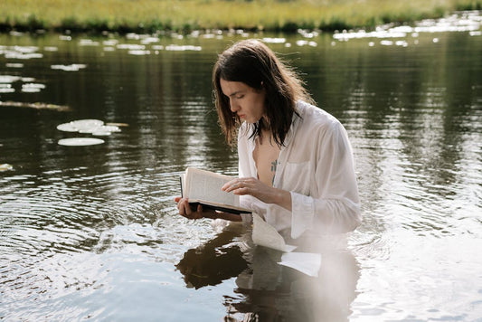 Person with tattoos in white shirt reads a book while standing in peaceful waters, creating a serene scene.