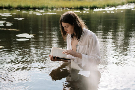 Person with tattoos in white shirt reads a book while standing in peaceful waters, creating a serene scene.