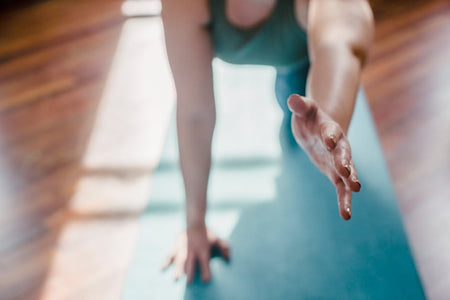 Person practicing yoga on a mat indoors, reaching forward in natural sunlight.