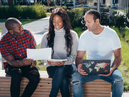 Three adults having a casual outdoor meeting discussing documents on a bench.