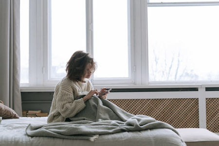 A woman in a knitted sweater using her smartphone while sitting on a bed with a blanket.