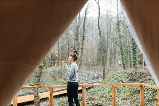 Woman sipping coffee in a serene forest setting, captured from a tent's perspective.