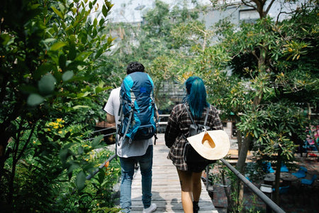 A couple with backpacks stroll on a wooden bridge surrounded by lush greenery, enjoying nature's tranquility.