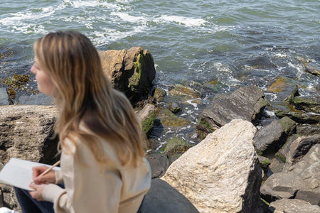 A woman sitting on rocks by the sea, writing in a notebook, enjoying a peaceful moment.